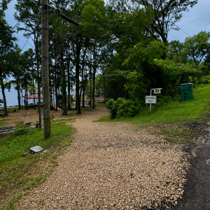 #image_title Gravel path through a wooded lakeside area with a power pole, trees, and an 'Entrance Only' sign near a green trash bin on a grassy slope.
