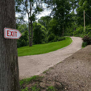 #image_title Red 'EXIT' sign attached to a tree beside a winding paved park path with lush green grass and trees nearby, daytime.