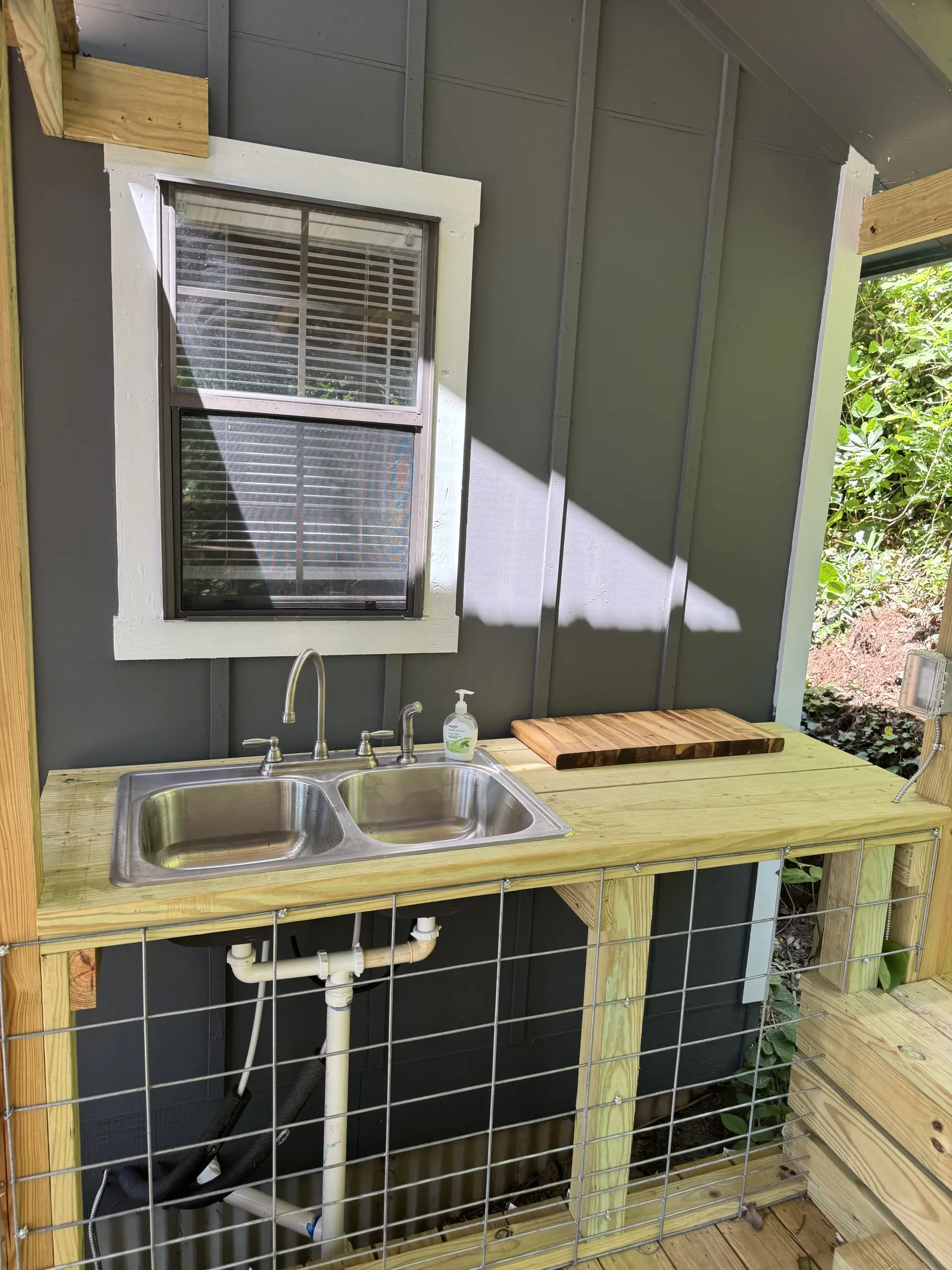 #image_title Outdoor kitchen setup with double-basin stainless steel sink, wooden counter, cutting board, and hand soap bottle against a gray siding wall with a window nearby