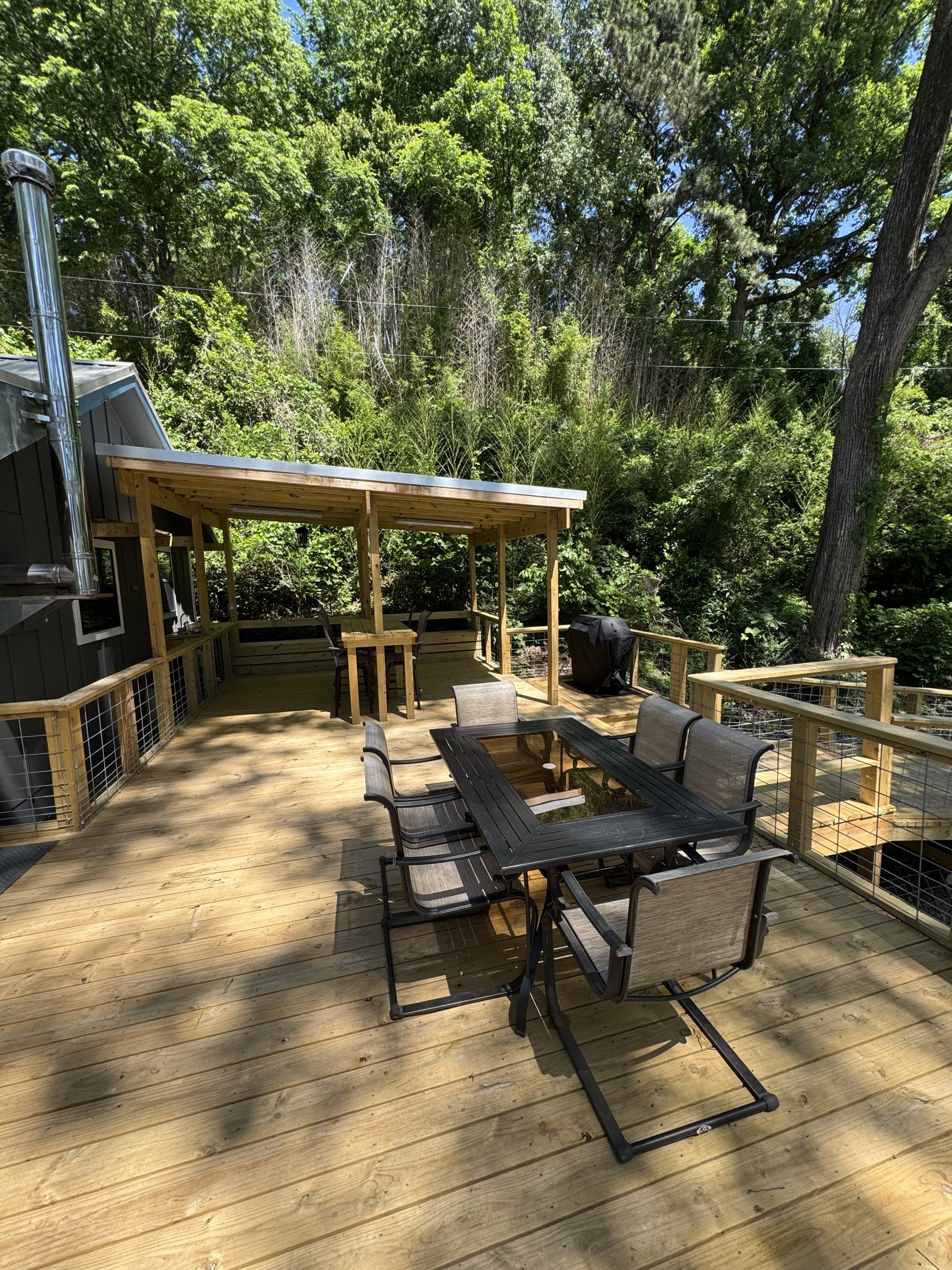 Wooden deck with outdoor dining table and chairs, railing, and a tree-filled backyard beyond a covered seating area.