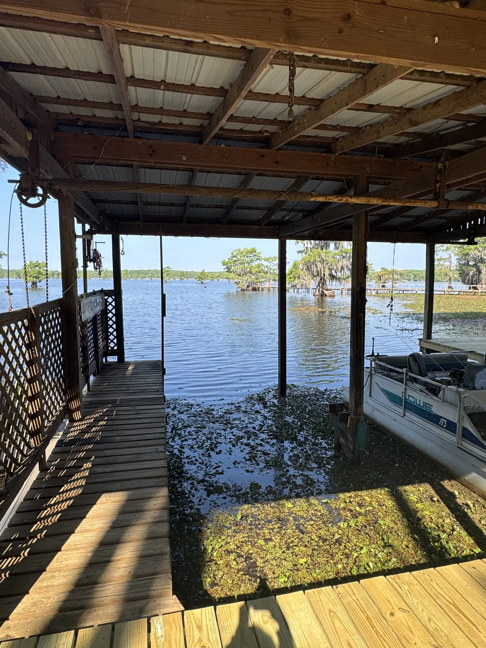 #image_title Wooden dock under a shed extends over a calm lake, with a small boat moored at the right and trees in the distance.