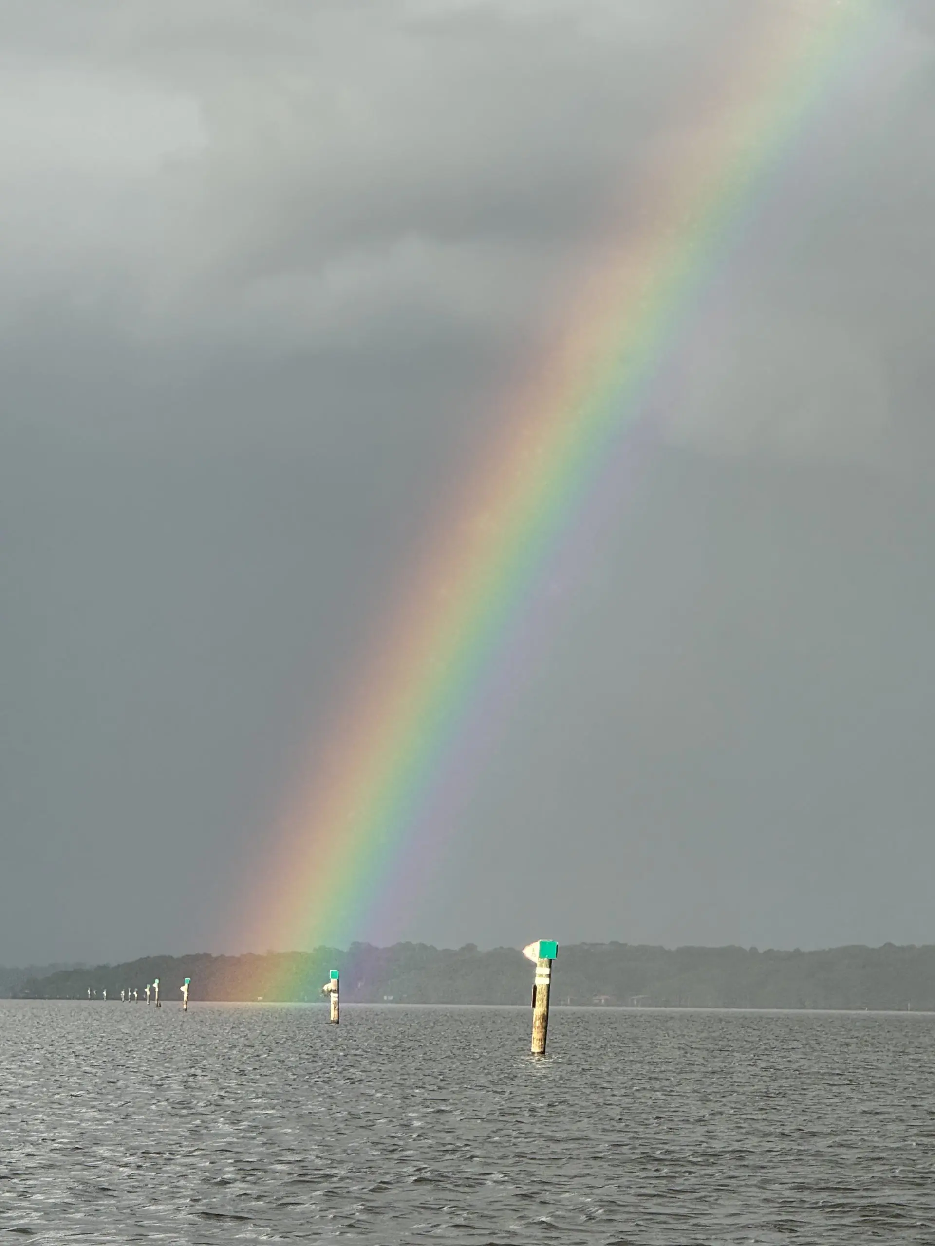 #image_title Rainbow arching over a cloudy sky above a calm body of water with wooden posts in the foreground.
